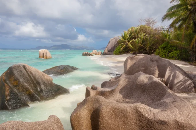 Playa con rocas grandes, agua turquesa y palmeras bajo un cielo nublado.